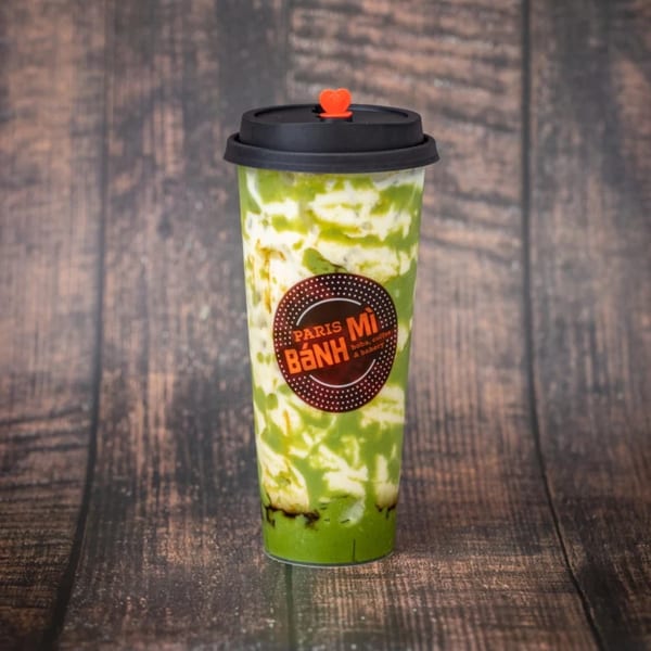 Paris Bánh Mì matcha drink with cream swirl and a heart-shaped stopper on a wooden background.