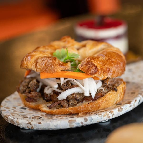 A Vietnamese croissant sandwich with beef, pickled carrots, onions, and cilantro on a speckled plate.