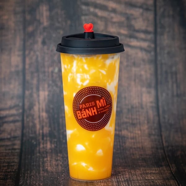 A Paris Bánh Mì mango drink with white swirls and a heart-shaped stopper on a wooden background.