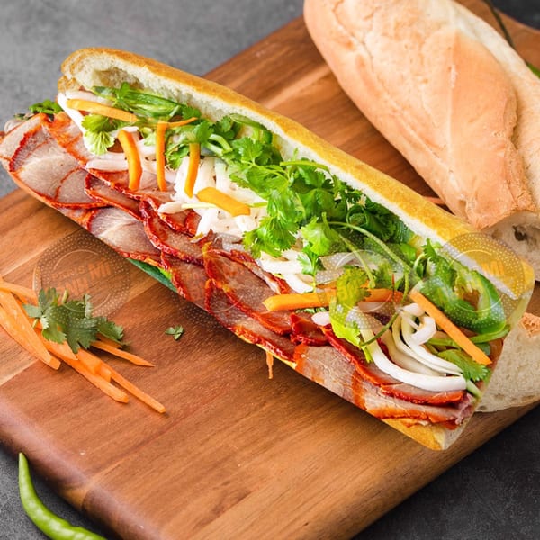 A Vietnamese bánh mì sandwich with sliced pork, fresh herbs, and pickled vegetables on a wooden board.