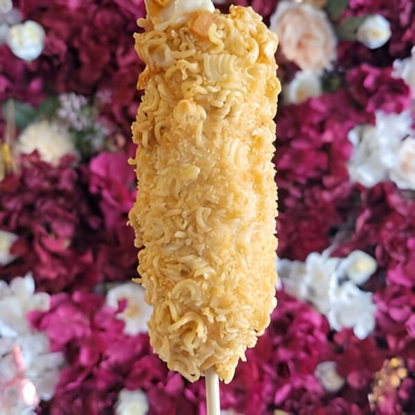 A crispy ramen-crusted corn dog on a stick, against a background of pink and white flowers.